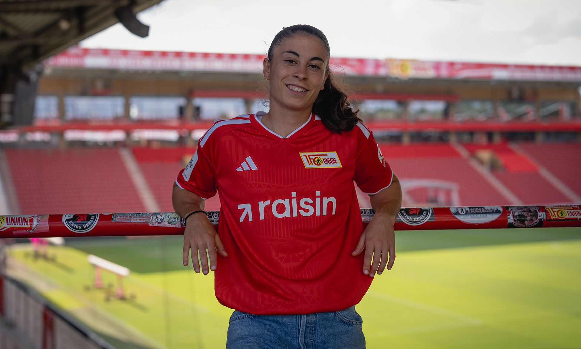 Eine junge Frau trägt ein rotes Fußballtrikot und lächelt, während sie an einem Geländer im Stadion lehnt.