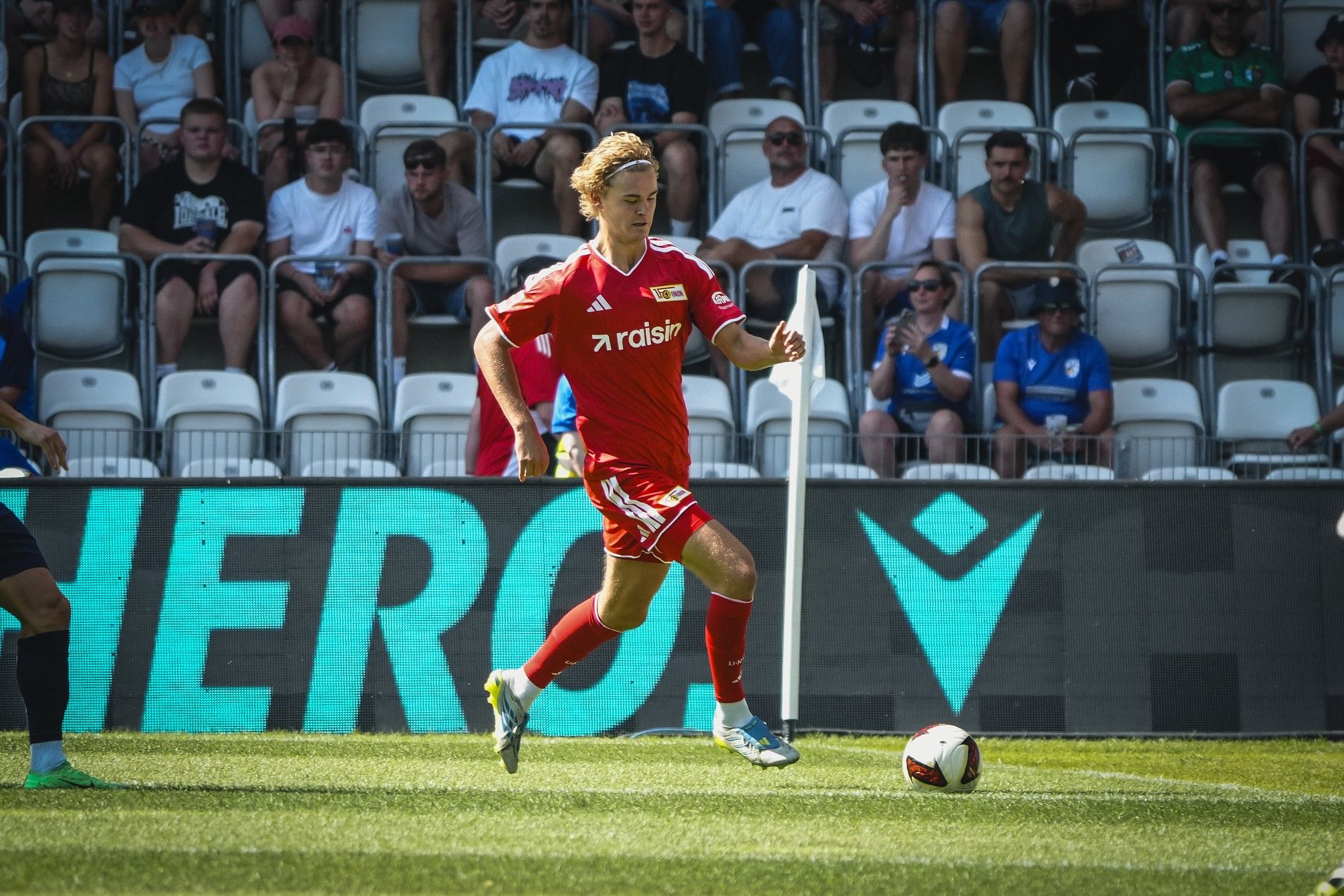 Ein junger Fußballspieler in einem roten Trikot sprintet mit dem Ball auf dem Spielfeld, während Zuschauer im Hintergrund zuschauen.