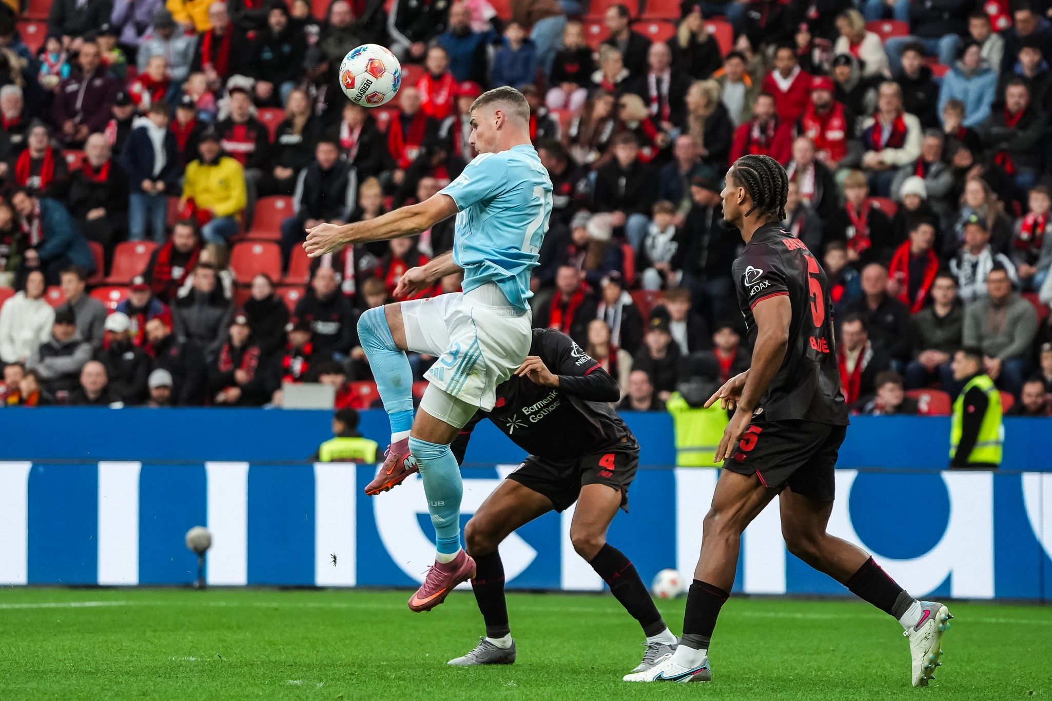 Ein Spieler in hellblauer Kleidung führt einen Luftball auf dem Fußballfeld aus, während zwei gegnerische Spieler versuchen, ihn zu stoppen.