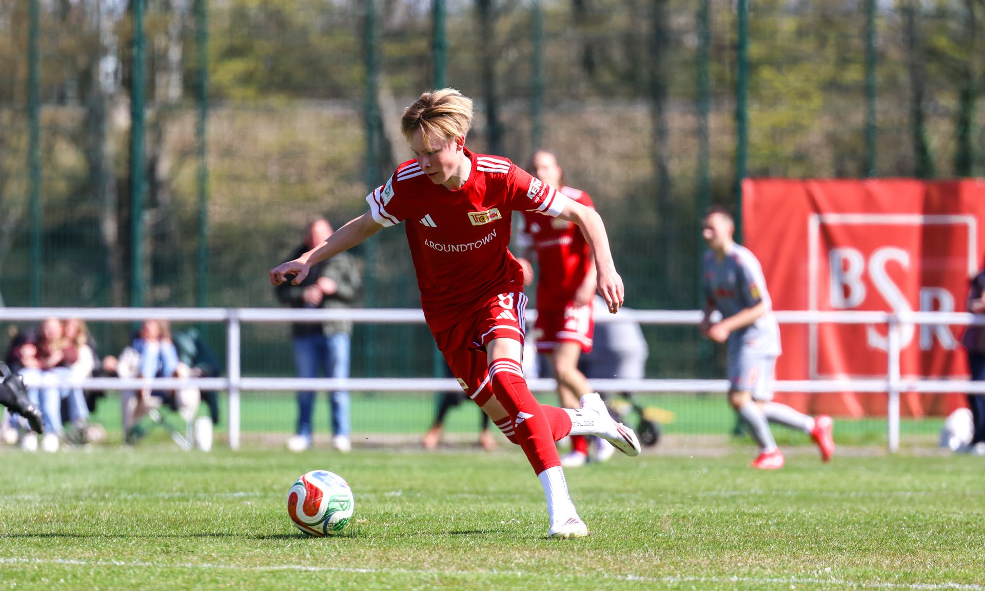 Ein junger Fußballspieler in rotem Trikot schießt einen Ball auf einem Sportplatz. Im Hintergrund sind weitere Spieler und Zuschauer zu sehen.