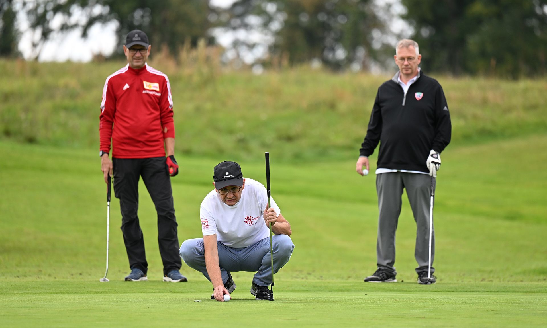 Drei Männer auf einem Golfplatz, einer sitzt auf dem Boden und richtet einen Golfball aus, die anderen beobachten.