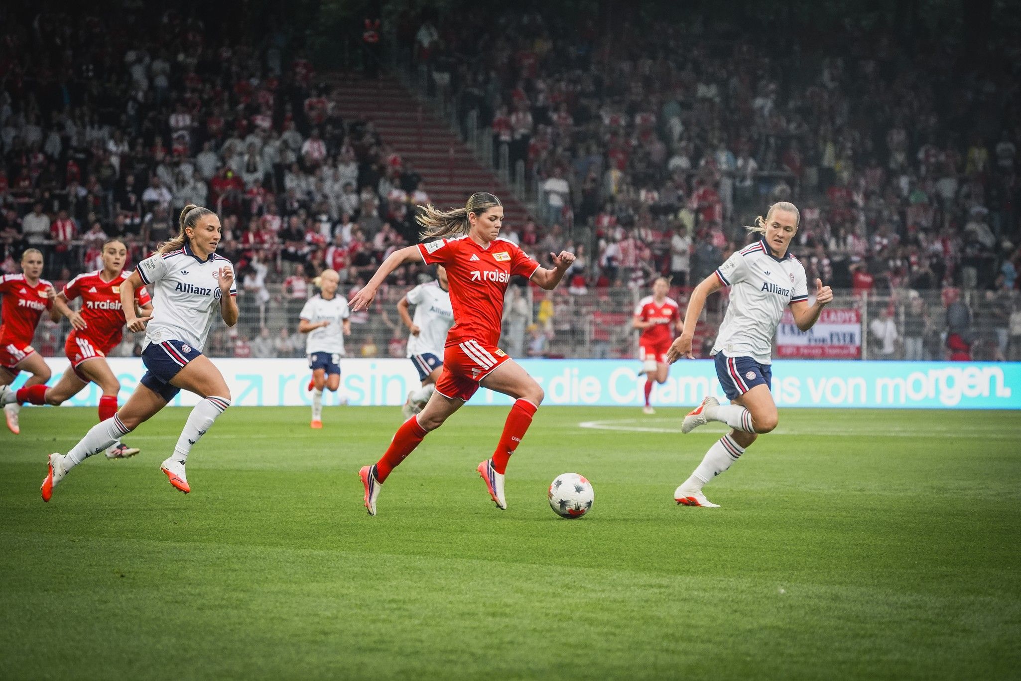 Eine Fußballspielszene mit zwei Teams, eine Spielerin in roter Kleidung und zwei in weißen Trikots, die um den Ball kämpfen.