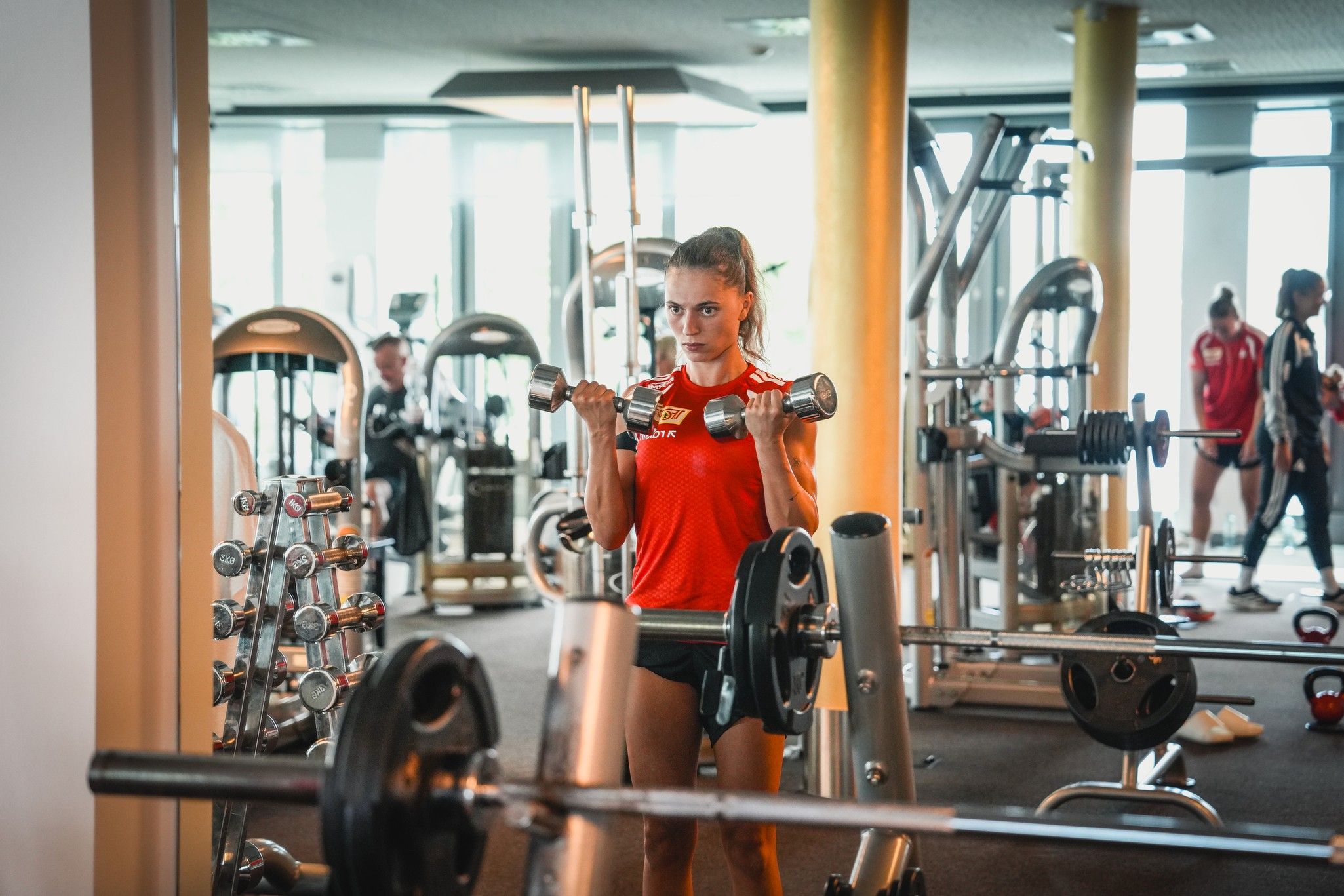Eine Sportlerin trainiert mit Hanteln in einem Fitnessstudio, umgeben von verschiedenen Fitnessgeräten.