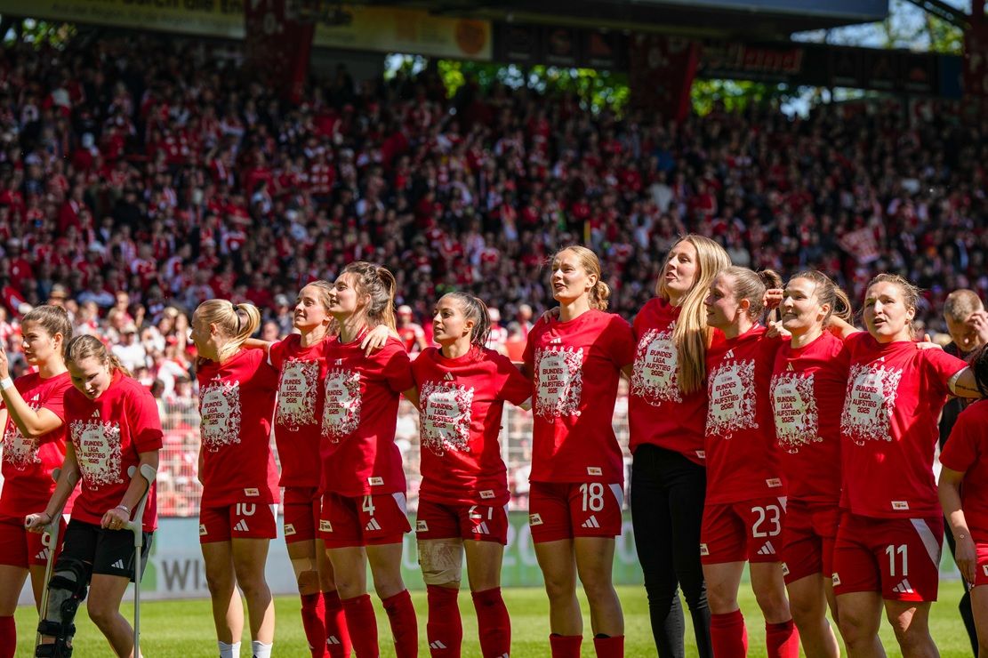 Frauenfußballmannschaft in roten Trikots singt zusammen im Stadion vor jubelndem Publikum.