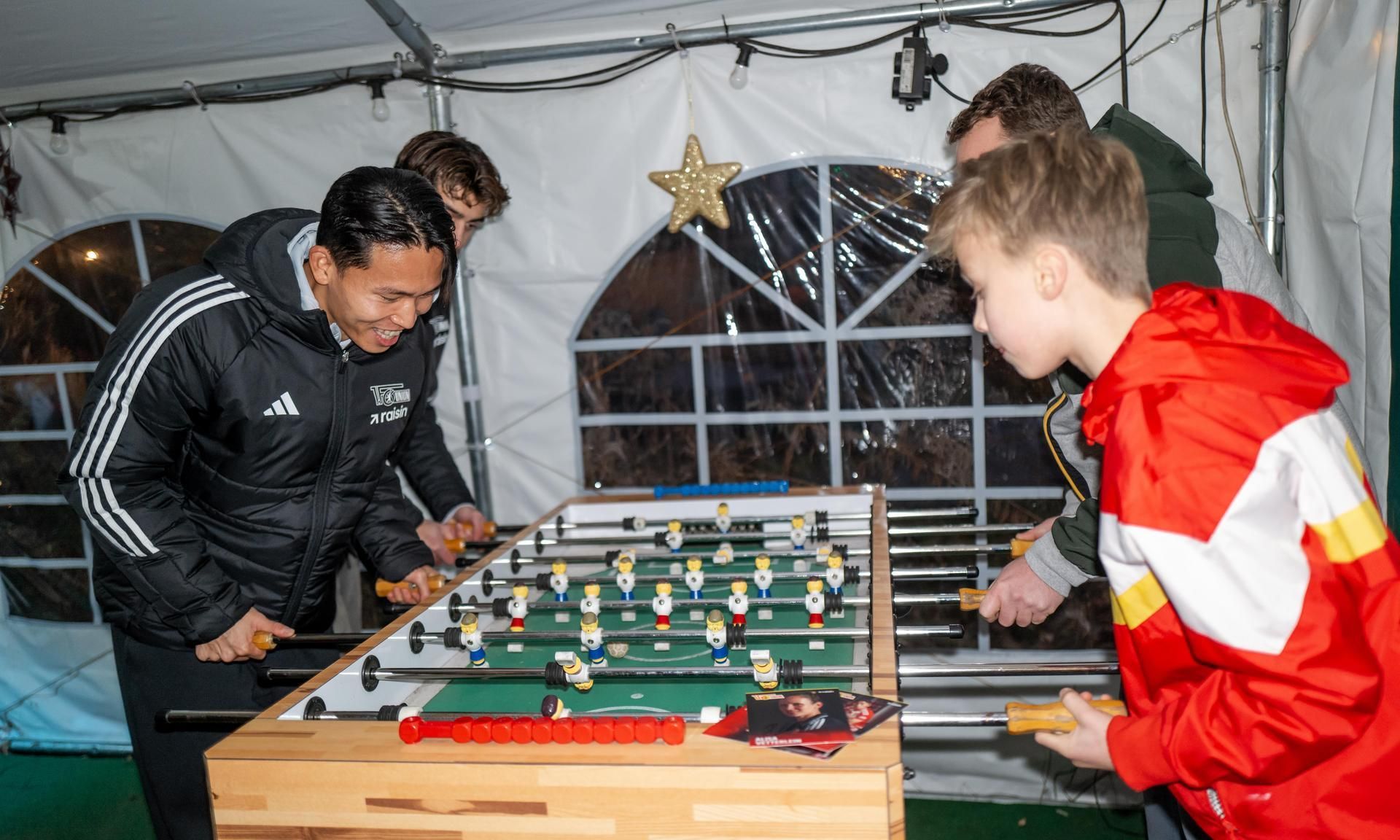 Zwei Erwachsene und ein Kind spielen Tischfußball in einem Zelt, während es draußen dunkel ist. Ein Stern ist dekorativ im Hintergrund.