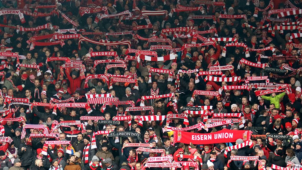 A large crowd of fans in red and white, holding scarves with "EISERN UNION" written on them and cheering.
