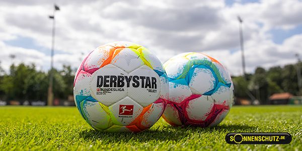 Two colorful footballs with the print "DERBYSTAR" are lying on green grass under a cloudy sky.