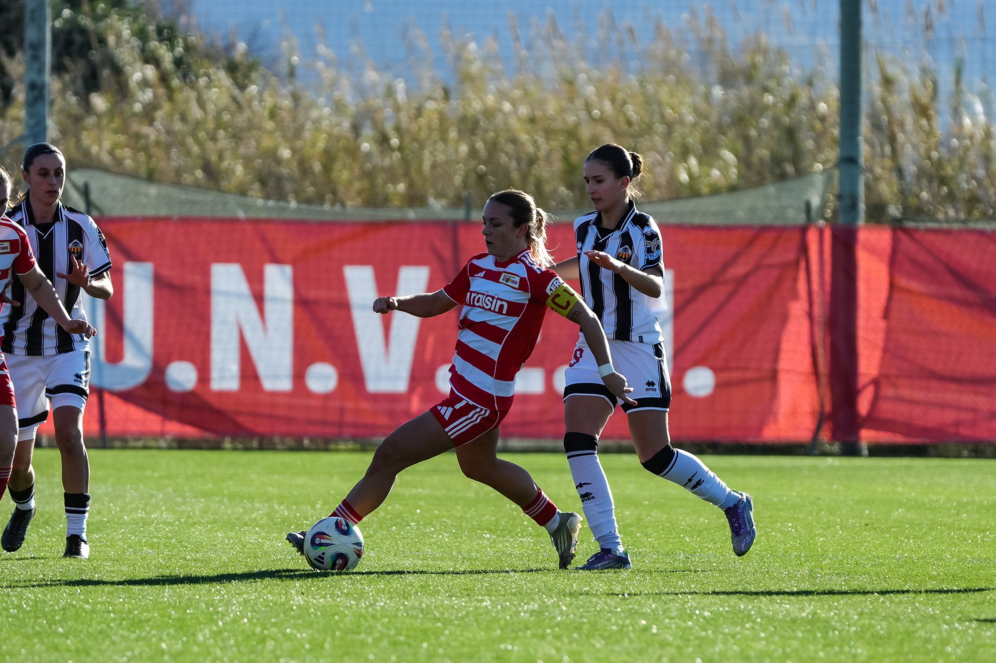 Eine Fußballspielerin in roter und weißer Uniform spielt den Ball, während eine Gegenspielerin in schwarz-weißer Kleidung sie verfolgt.