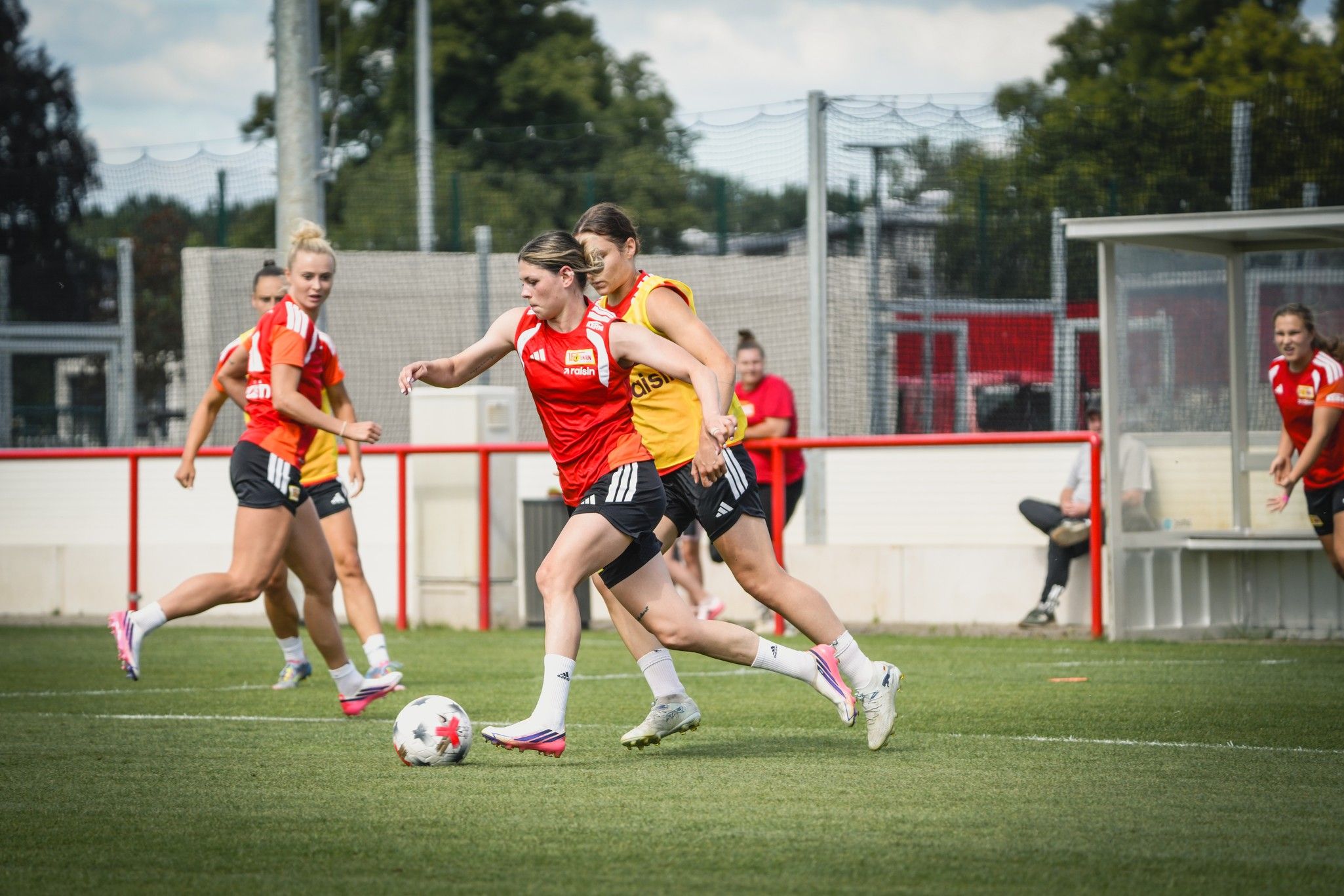 Frauenfußballtraining auf einem Rasenplatz, Spielerinnen in roten und gelben Trikots, mit einem Ball im Spiel.