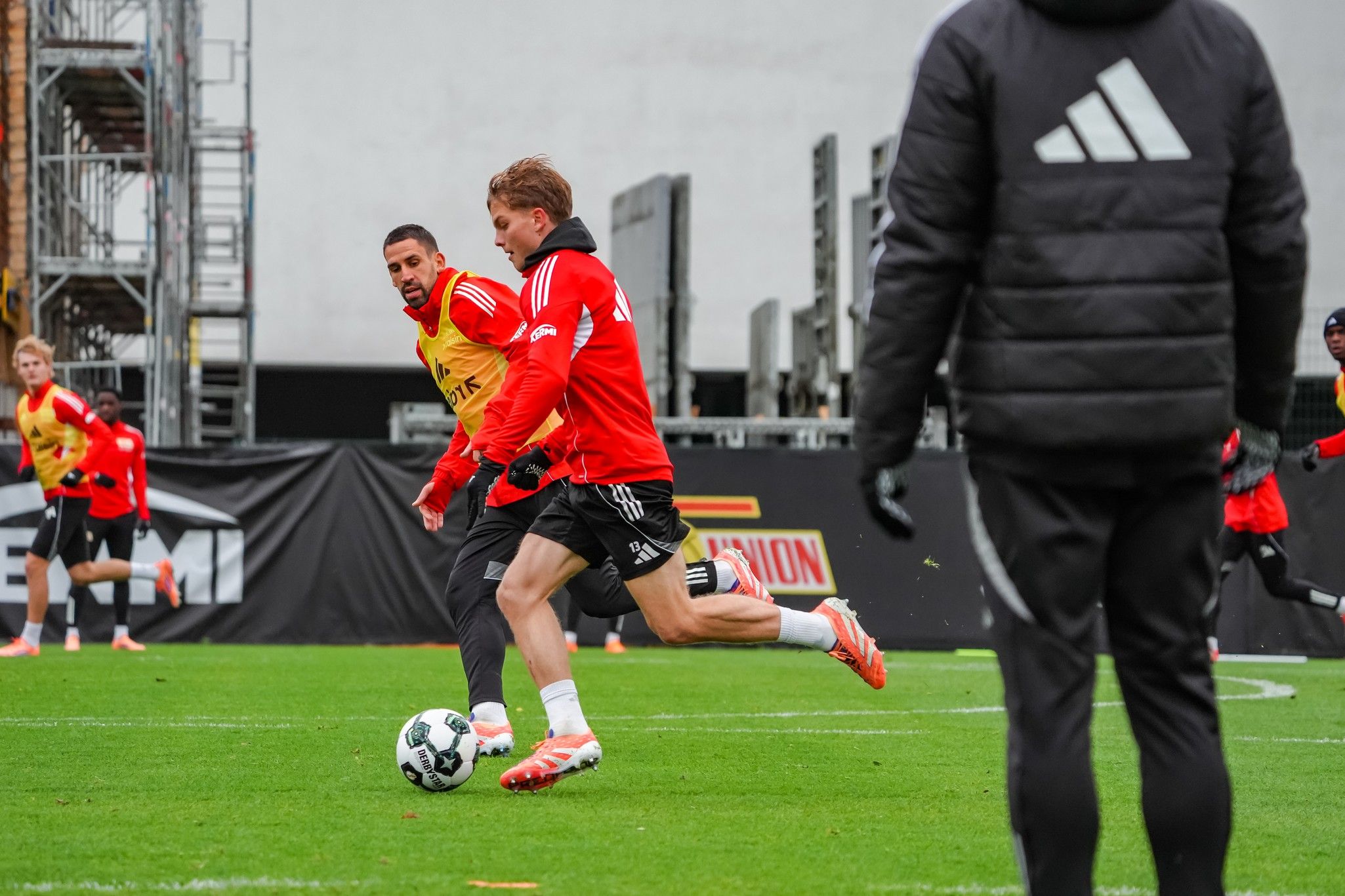 Zwei Fußballspieler in roten Trikots üben auf einem Trainingsfeld, während ein Trainer im Hintergrund zusieht.