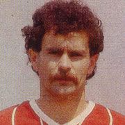 Ein Mann mit lockigem, braunem Haar und einem Schnurrbart trägt ein rotes Fußballtrikot.