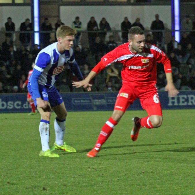 Zwei Fußballspieler stehen sich auf dem Spielfeld gegenüber, einer läuft in Richtung Tor, während der andere ihn verfolgt.