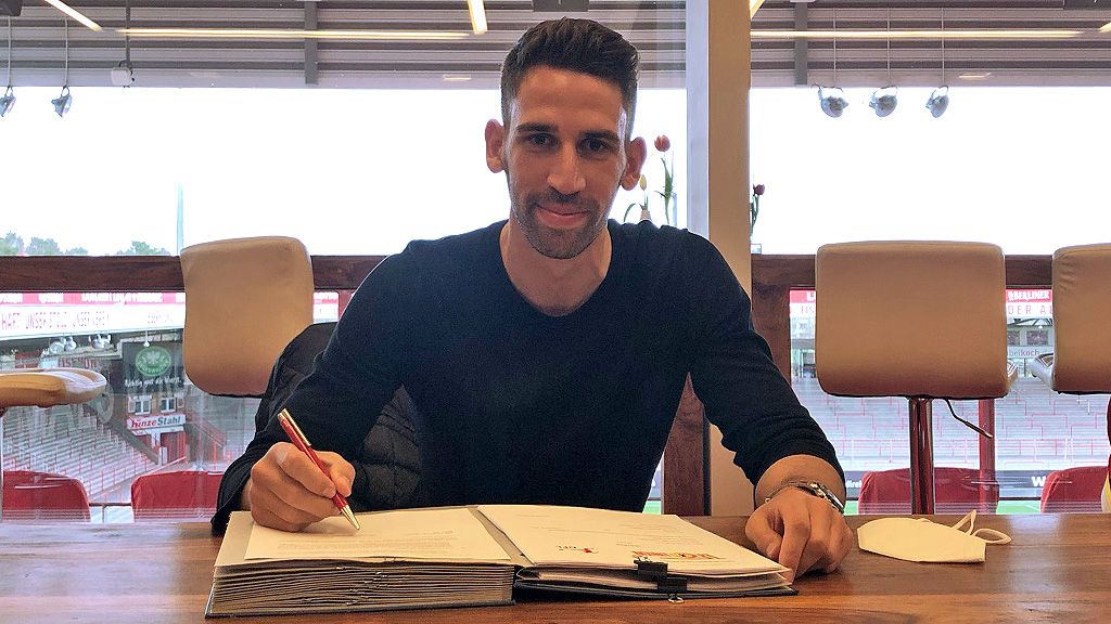 Ein Mann sitzt an einem Tisch und schreibt in ein Buch, während er in die Kamera lächelt. Pferde sind im Hintergrund sichtbar.