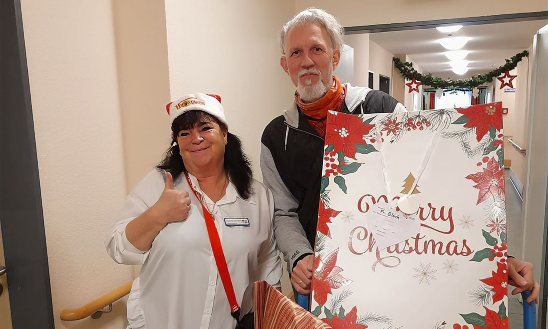 Eine Frau mit Weihnachtsmütze und Daumen hoch steht neben einem Mann, der ein großes Geschenk mit Weihnachtsmotiv hält.