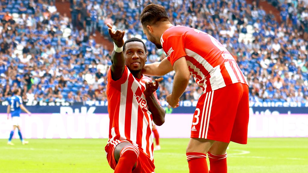 Ein Fußballspieler mit roten und weißen Kleidern sitzt auf dem Gras. Ein Kollege steht neben ihm. Sie freuen sich.