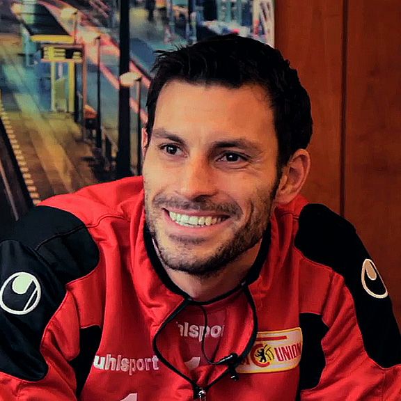 Ein lächelnder Mann in sportlicher Kleidung sitzt vor einem Hintergrund mit urbanem Motiv.