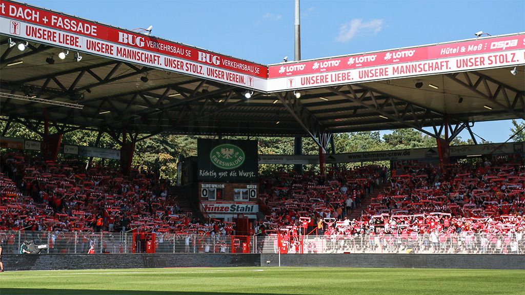 Vollbesetzte Tribüne in einem Stadion mit Fans, die rote Schals halten, unter blauem Himmel und Sonnenschein.