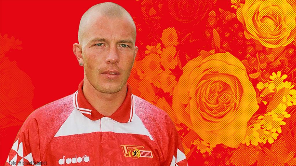 Ein junger Mann mit kurzem Haar trägt ein rot-weißes Fußballtrikot und steht vor einem bunten, floralen Hintergrund.