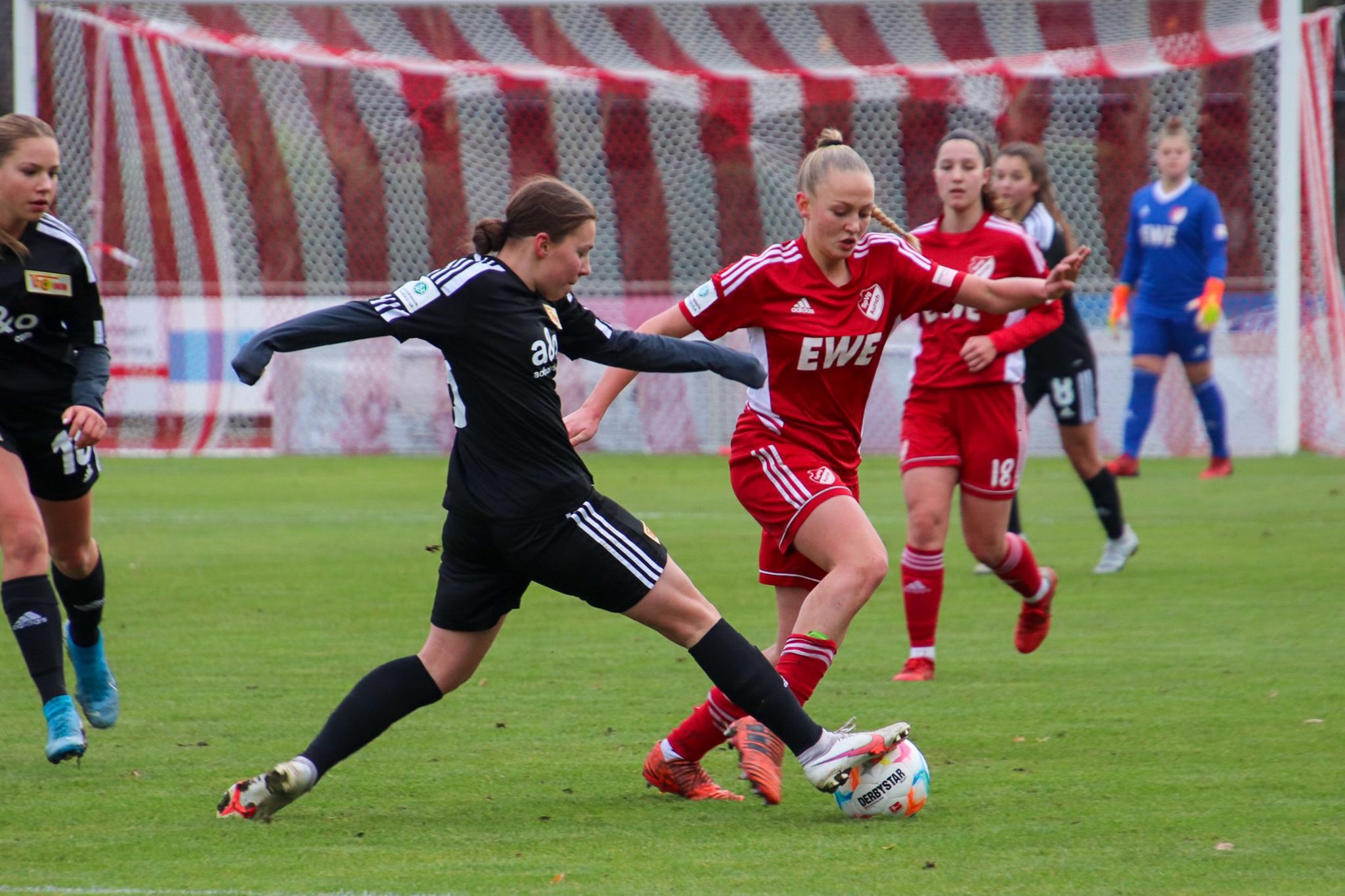 Eine Spielerin in schwarzer Trikot versucht, den Ball von einer Spielerin in rotem Trikot zu trennen. Im Hintergrund sind weitere Spielerinnen.