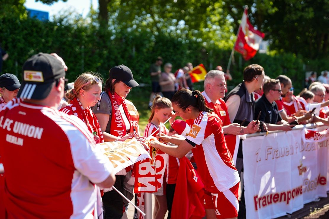 Gruppierung von Fans in roten Trikots, die Banner halten und auf ein Sportereignis warten. Sonnenschein und festliche Atmosphäre.