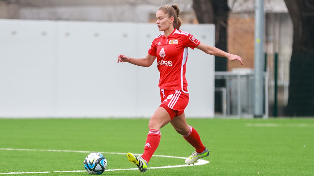 A female soccer player in red sportswear kicks the ball on a green artificial turf field.