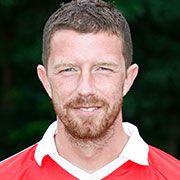 Porträt eines lächelnden Mannes mit Bart in rotem Trikot vor unscharfem grünem Hintergrund.