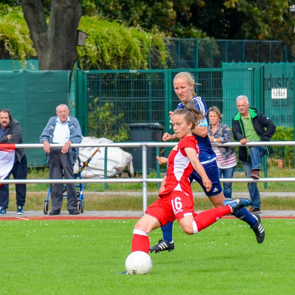 Eine Spielerin in Rot greift nach dem Ball, während eine Spielerin in Blau sie von hinten angreift. Zuschauer beobachten das Spiel im Hintergrund.