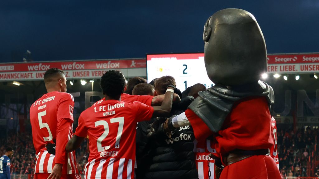 Players of 1. FC Union Berlin celebrate a goal surrounded by fans and a mascot in the stadium.