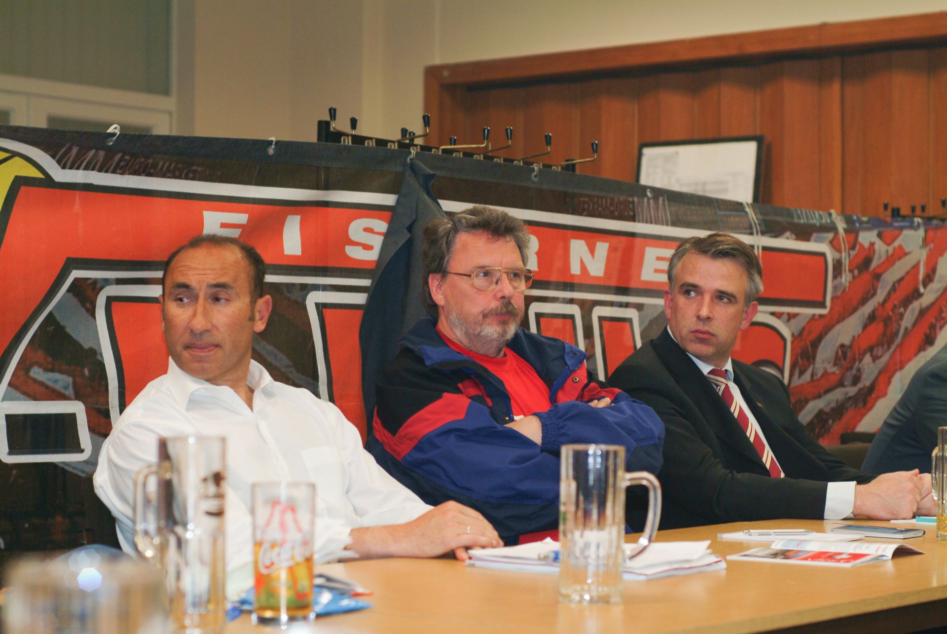 Drei Männer in formeller Kleidung sitzen an einem Tisch, während im Hintergrund ein Banner mit dem Wort "FISCHER" sichtbar ist.
