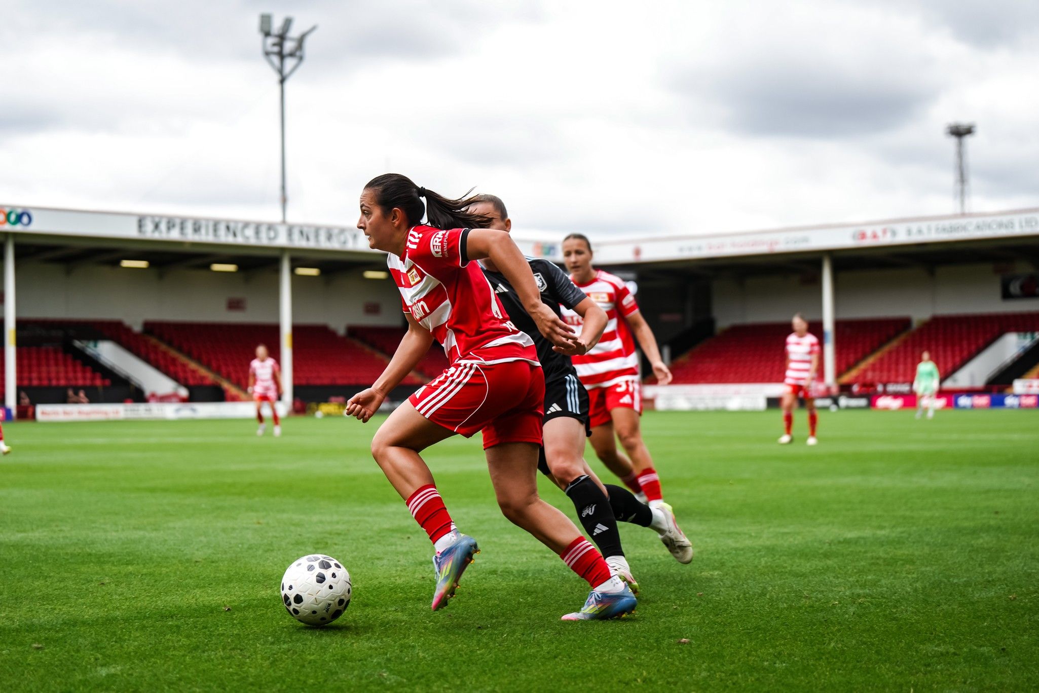 Eine Spielerin in rotem Trikot dribbelt den Ball auf einem Fußballfeld, während eine Gegenspielerin sie verfolgt.
