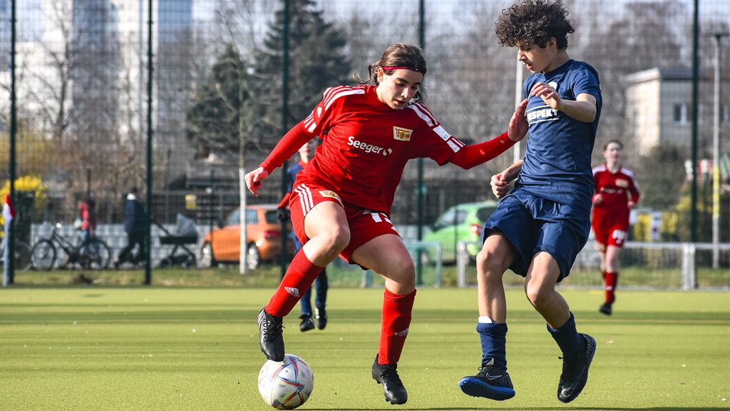 Ein Mädchen in rotem Fußballtrikot dribbelt den Ball, während ein Junge in blauem Trikot versucht, ihn zu stoppen.