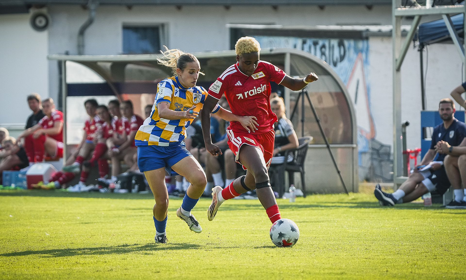 Zwei Fußballerinnen im Wettlauf um den Ball auf einem grünen Spielfeld, während Zuschauer auf der Bank zuschauen.
