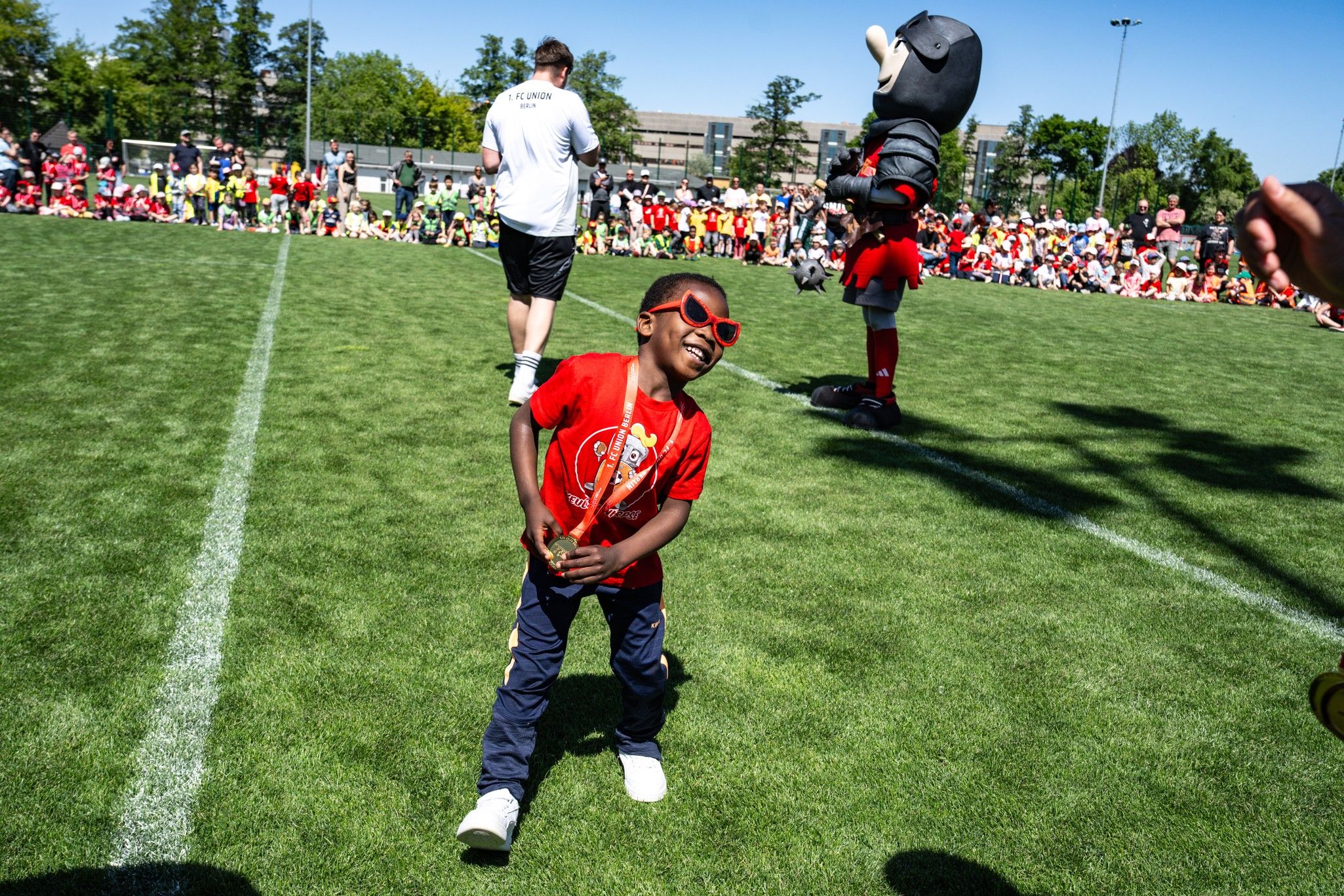 Ein fröhlicher Junge in Sonnenbrille hält eine kleine Trophäe und lacht auf einem Sportfeld, umgeben von Kindern und Figuren im Kostüm.