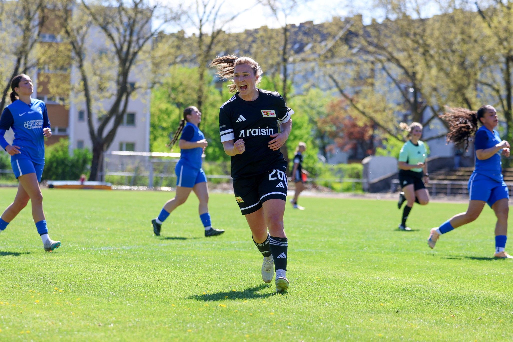 Eine Spielerin in schwarzer Ausrüstung jubelt auf dem Fußballplatz, während andere Spielerinnen im Hintergrund aktiv sind.