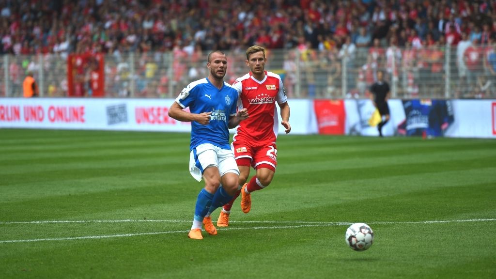 Zwei Fußballspieler in blauen und roten Trikots stehen auf einem Spielfeld während eines Spiels.