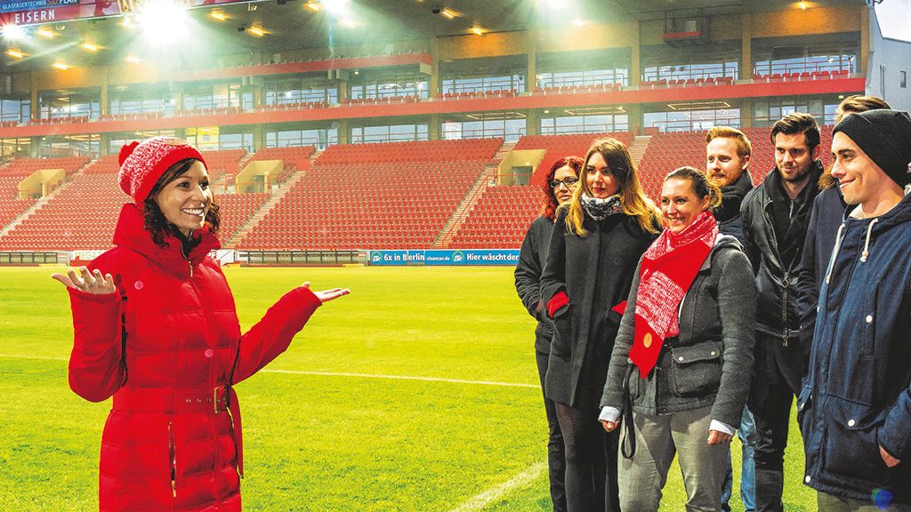 Eine Gruppe von Menschen steht auf einem Fußballfeld, während eine Frau in roter Jacke ihnen etwas erklärt.