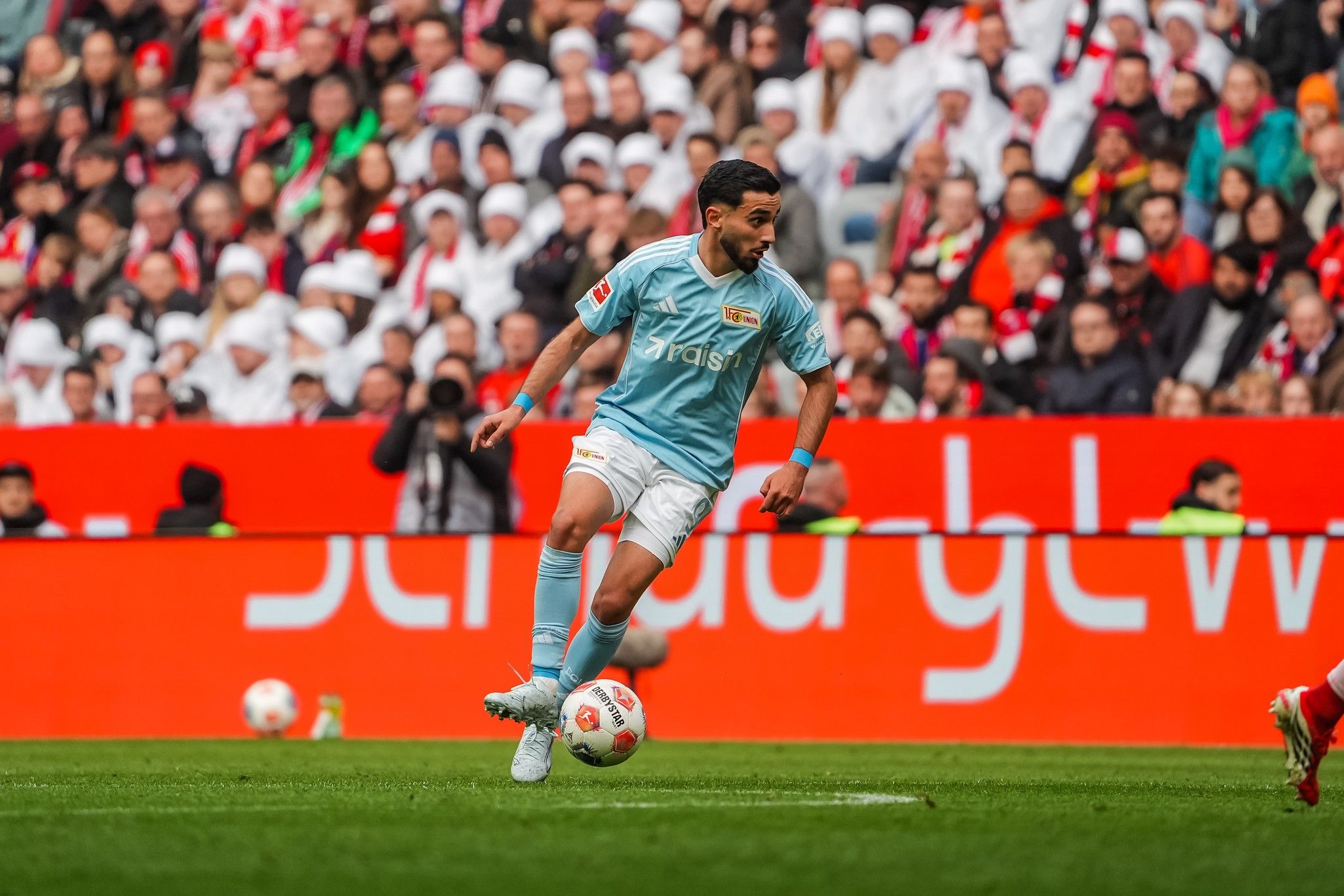 Spieler in hellblauer Fußballausrüstung dribbelt den Ball auf dem Spielfeld während eines Spiels mit Zuschauern im Hintergrund.