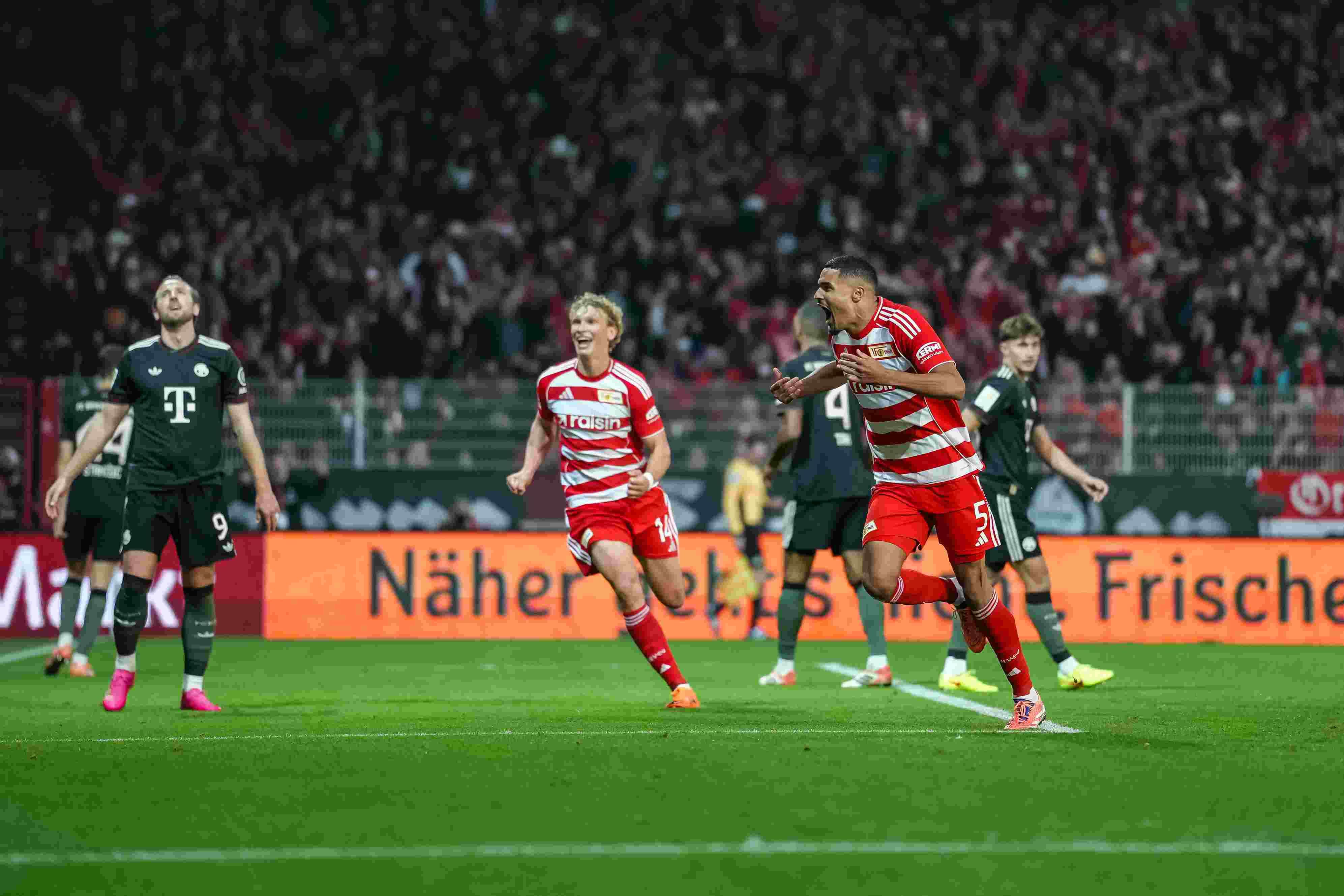 Ein Spieler freut sich über ein Tor. Seine Teamkollegen jubeln. Im Hintergrund sieht man das andere Team. Es gibt eine gute Stimmung im Stadion.