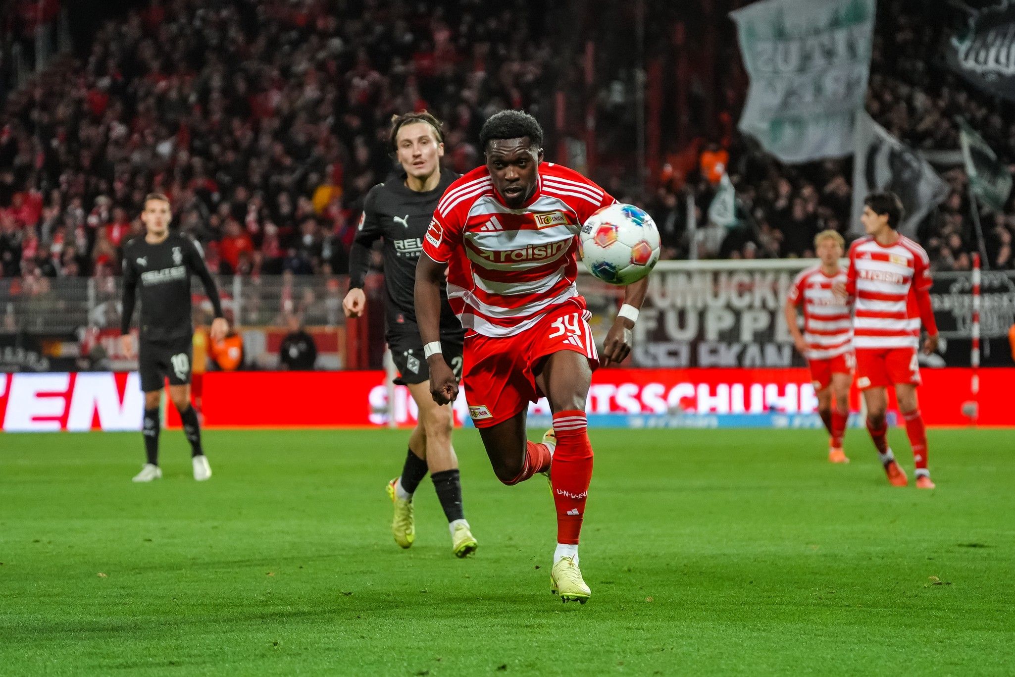 Ein Fußballspieler in roten Trikots sprintet mit dem Ball, während ein Gegner ihn verfolgt. Publikum im Hintergrund.