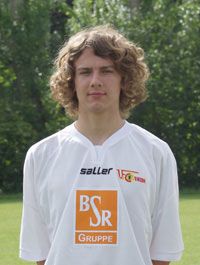 Junger Mann mit lockigem Haar, trägt ein Fußballtrikot mit Sponsorenlogo, steht im Freien vor einer grünen Kulisse.