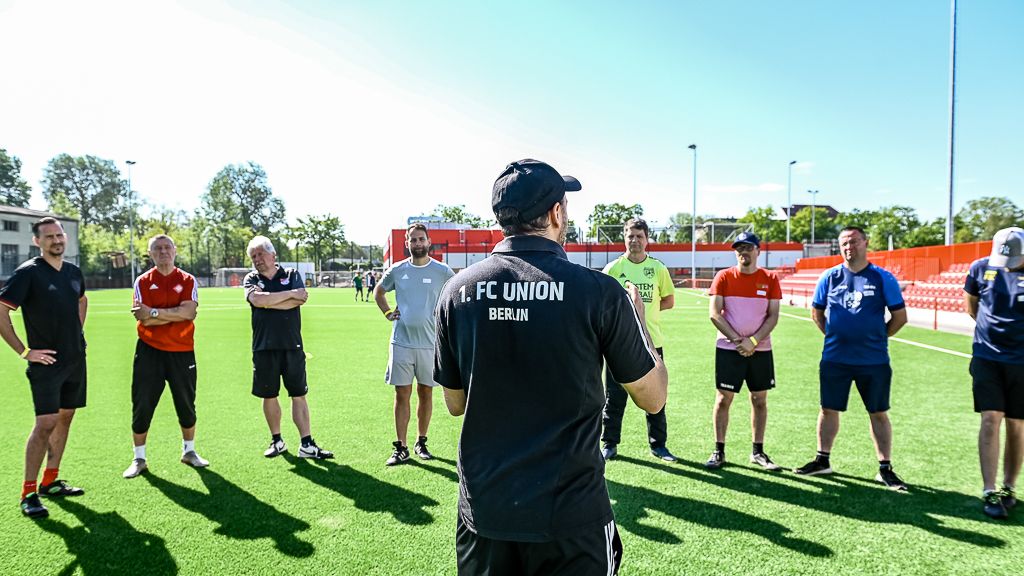 Trainer steht vor einer Gruppe von Fußballtrainern auf einem Trainingsplatz mit Kunstrasen.