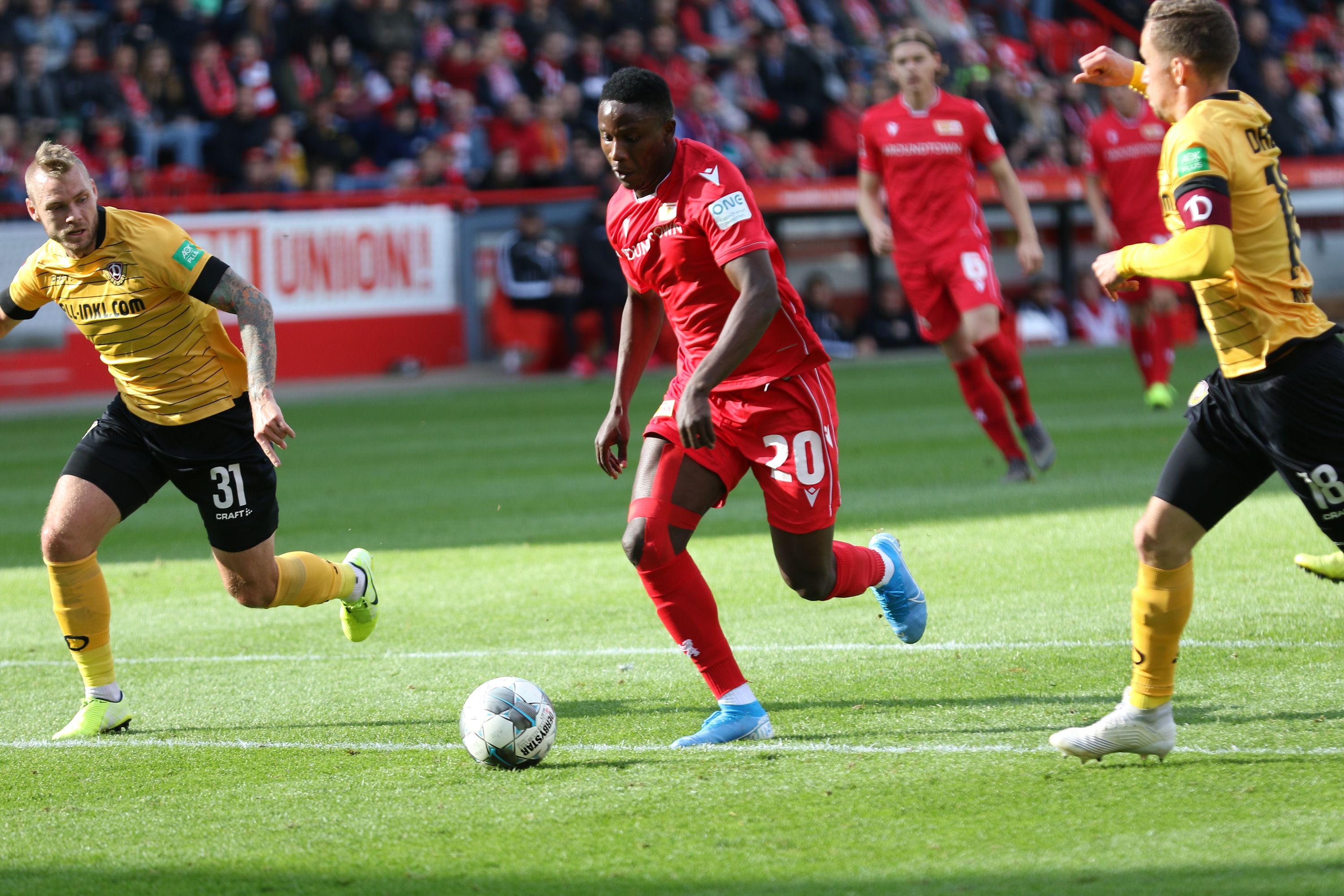 Ein Fußballspieler in rotem Trikot dribbelt den Ball, während zwei gegnerische Spieler ihn verfolgen.