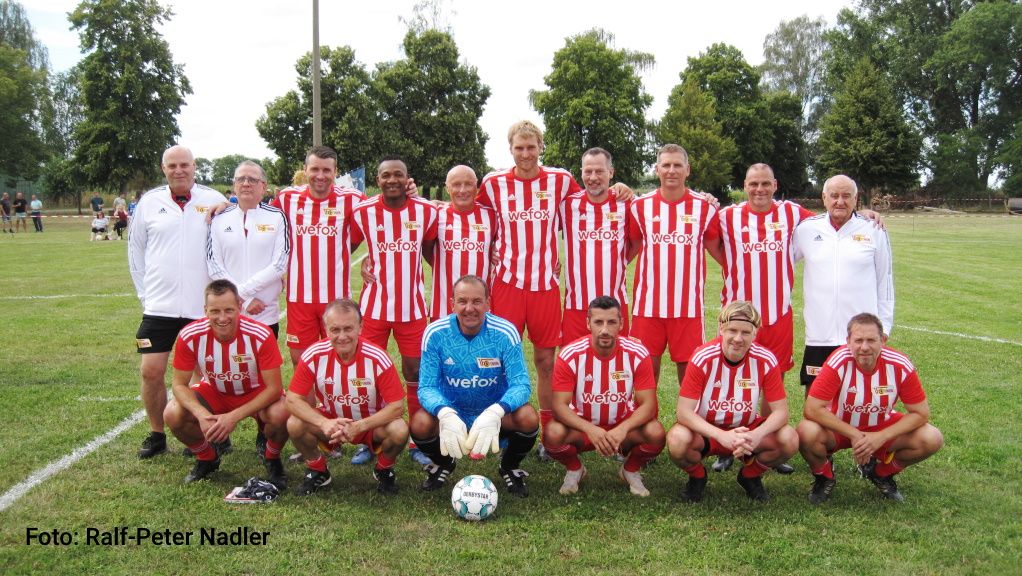 Eine Gruppenfoto von 12 Fußballspielern in rot-weißen Trikots, stehen auf einem Rasenfeld, umgeben von Bäumen.