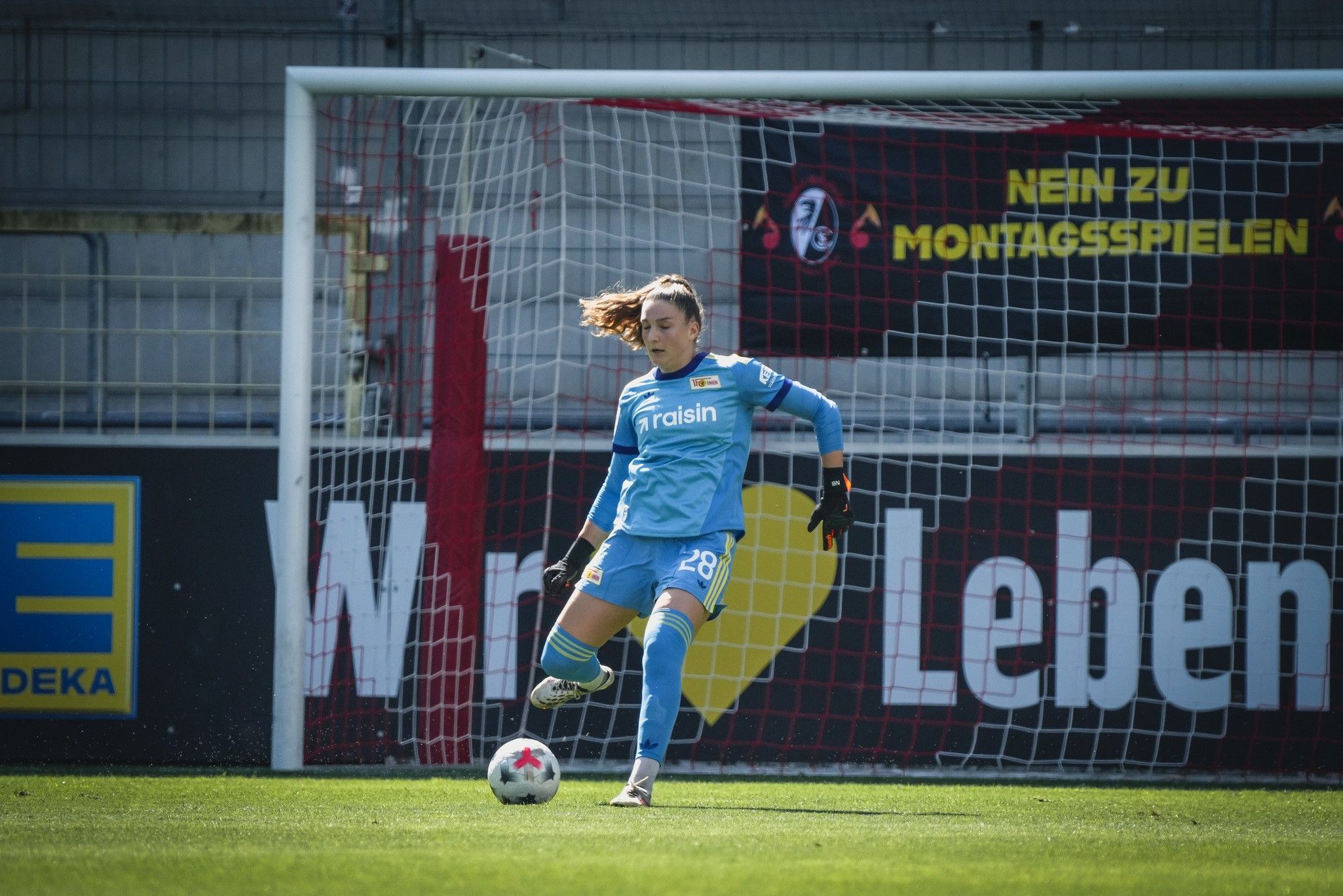 Torhüterin in blauer Ausrüstung steht im Tor, bereit, den Ball zu spielen. Im Hintergrund ein Fußballtor und Werbebanner.