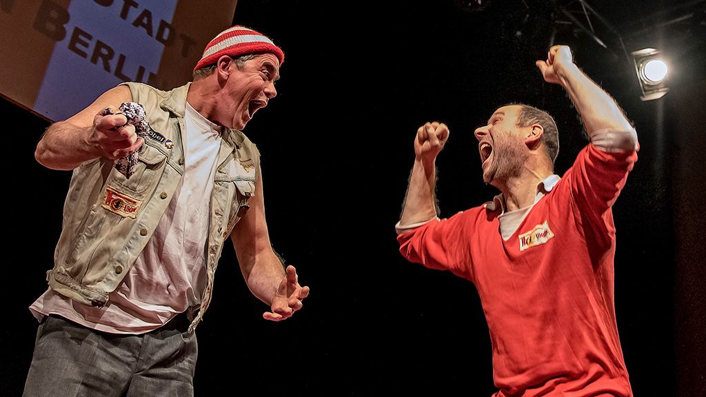 Zwei Männer auf der Bühne, einer in einem ärmellosen Hemd und der andere in einem roten Pullover, zeigen emotionale Gesten.
