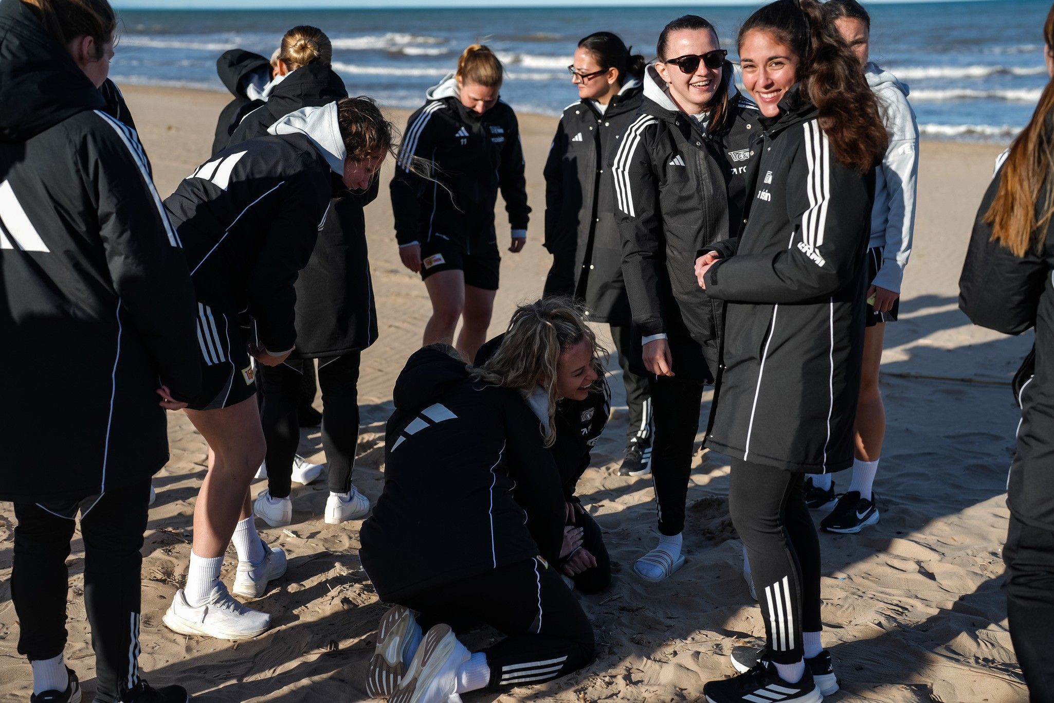 Gruppe von Jugendlichen in sportlicher Kleidung am Strand, einige lachen und interagieren, während andere im Sand spielen.