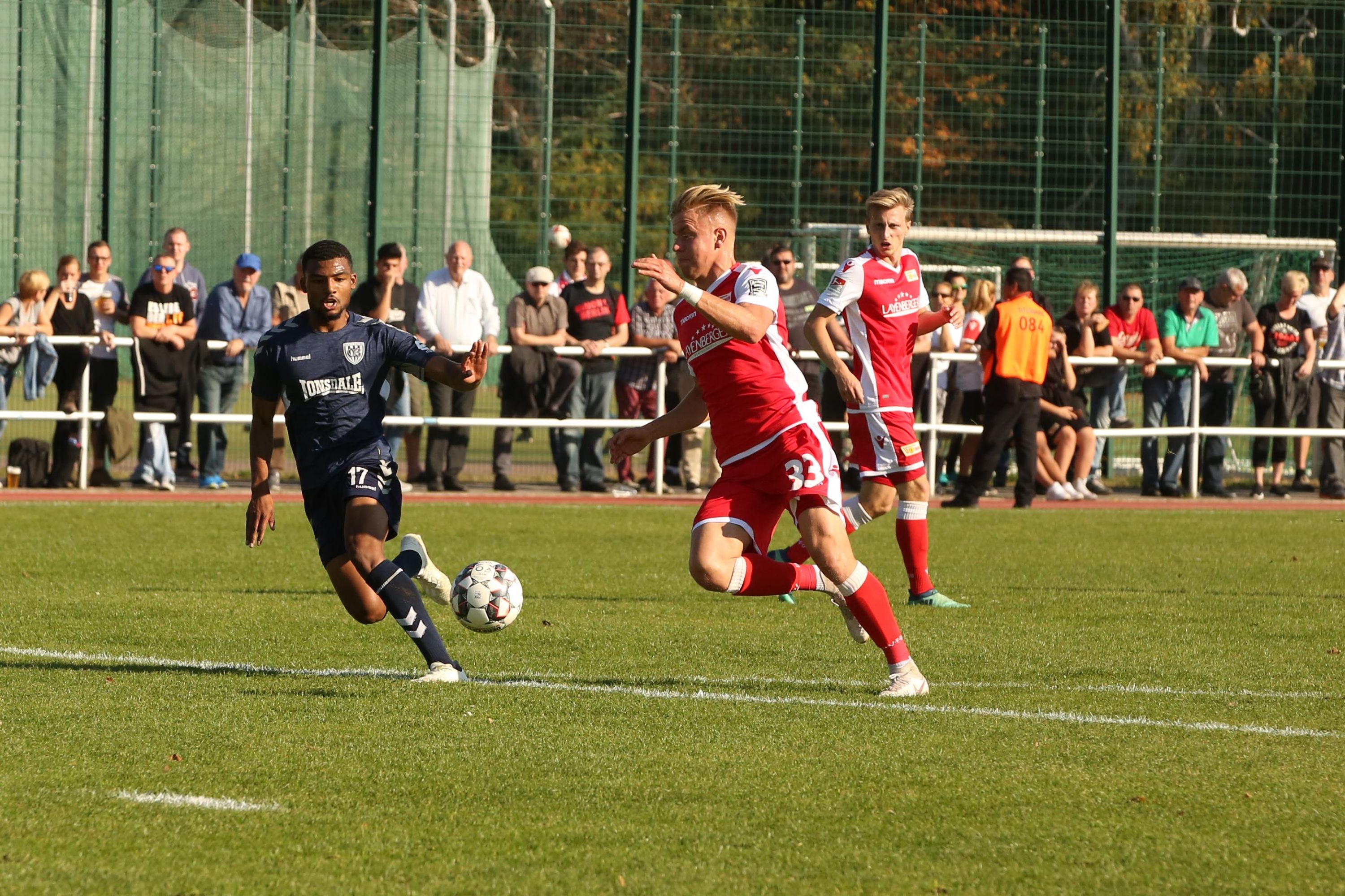 Two football players in uniform chase the ball on a sports field while spectators watch in the background.