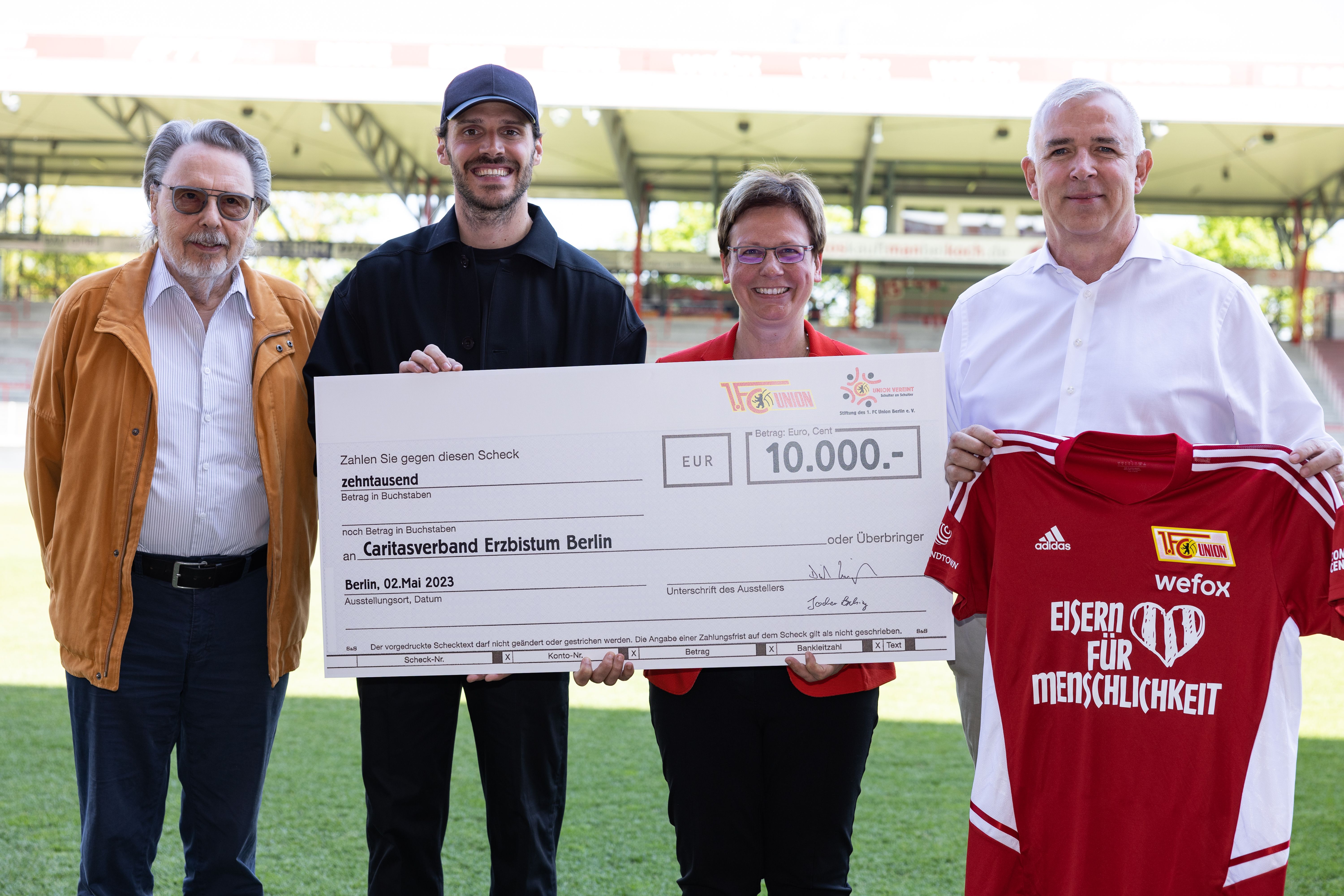 Vier Personen stehen auf einem Sportplatz. Sie halten einen großen Scheck. Der Scheck ist über 10.000 Euro. Der Scheck ist für einen Verein.