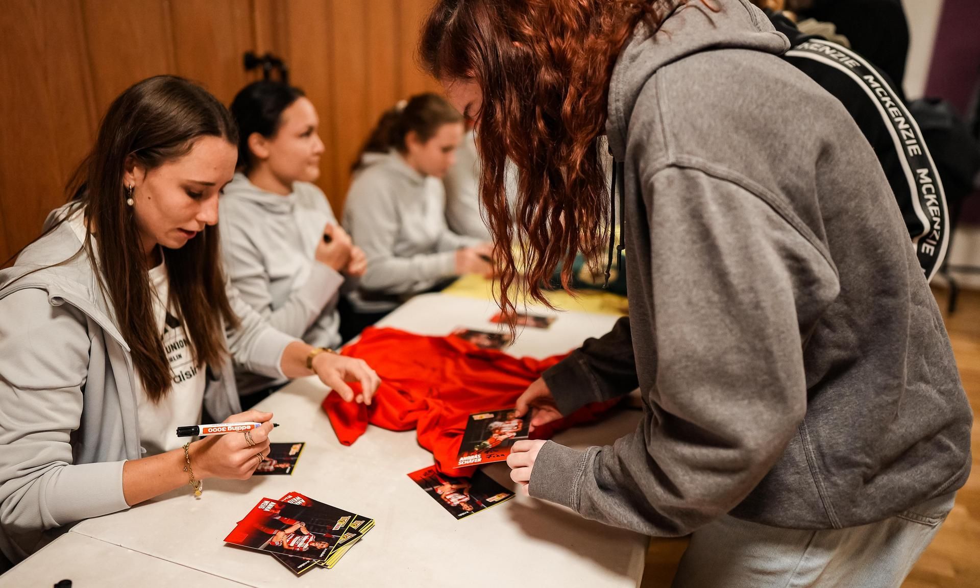 Eine Gruppe von Frauen sitzt an einem Tisch und signiert Autogrammkarten für eine wartende Person.