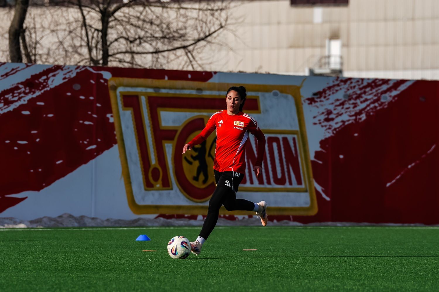 Eine Person in einem roten Sportshirt läuft mit einem Fußball auf einem grünen Spielfeld, im Hintergrund eine farbige Wand.